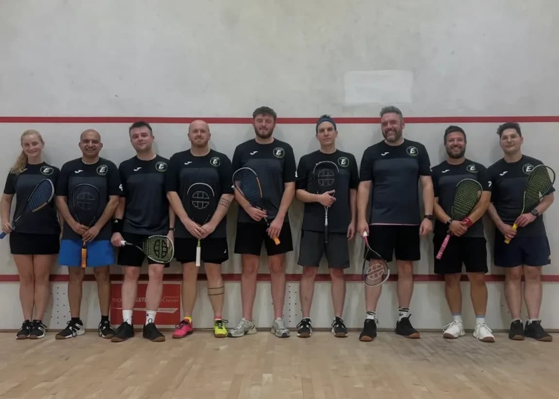 Nine Exton Park players in black club shirts, lined up against a squash court wall with their rackets